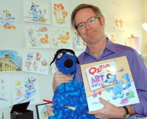 By Christina Corrales-Toy Surrounded by walls of his work in his Newcastle home, local illustrator Dana Sullivan poses with a puppet-version of Ozzie, the star of his first children’s book, ‘Ozzie and the Art Contest.’ 